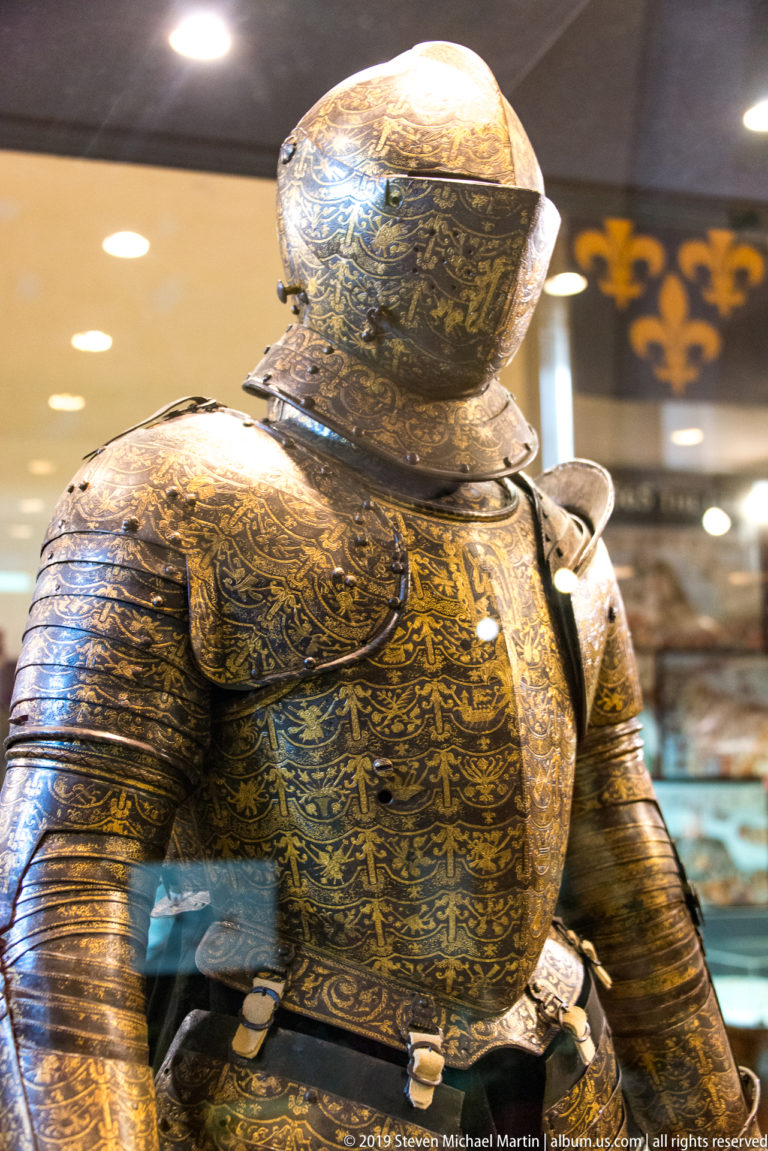 Close view of dark full plate armor densely decorated with gold-toned etched or inlaid patterns across helmet, chest, shoulders, and arms.