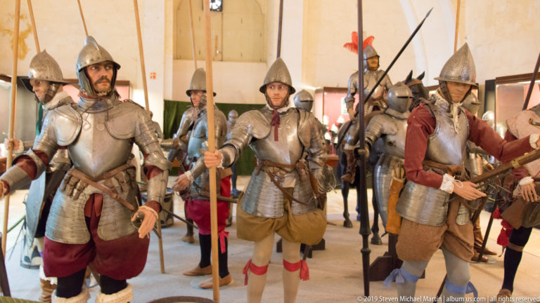 Group of armored soldier mannequins holding long pikes and polearms, wearing varied helmets, breastplates, and red or buff garments.