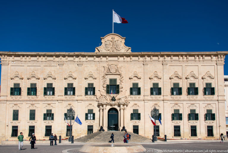 Wide frontal view of Auberge de Castille across the square, with symmetrical green-shuttered windows, central entrance, flags, and pedestrians.