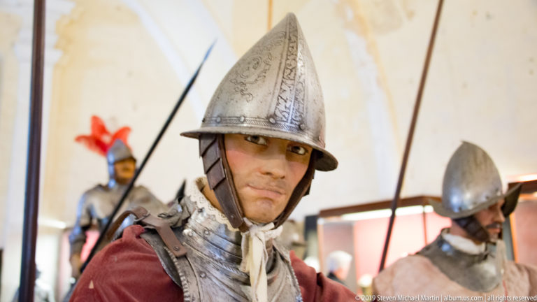 Close portrait of a helmeted soldier mannequin in polished breastplate and red clothing, with armored figures and weapons behind.