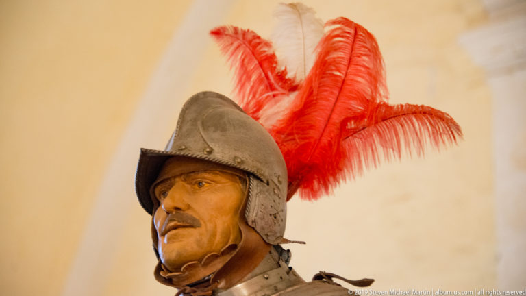 Close profile of an armored mannequin wearing a pointed metal helmet with neck protection and a large red feather plume.