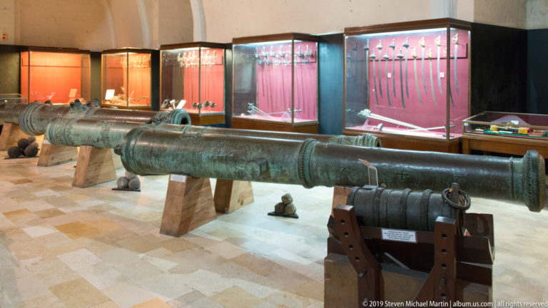 Large historic cannon barrels on wooden supports in a museum gallery, with red-lit cases of swords and smaller weapons behind.