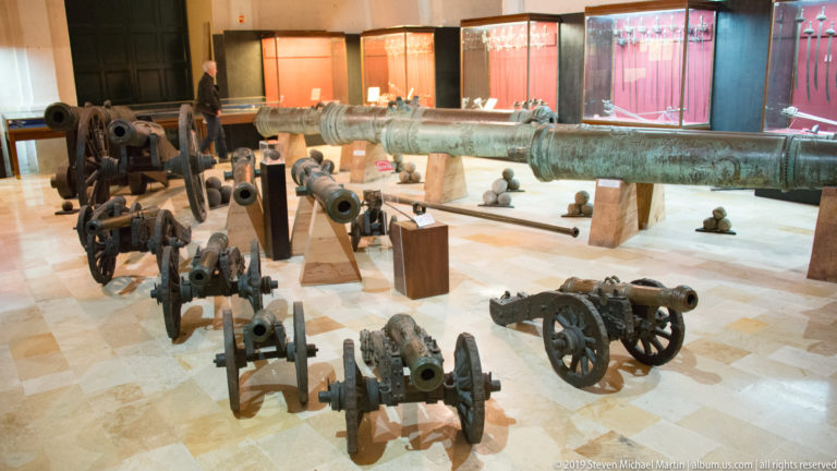Museum gallery displaying several historic wheeled cannon, a long bronze gun, cannonballs, and other artillery pieces.