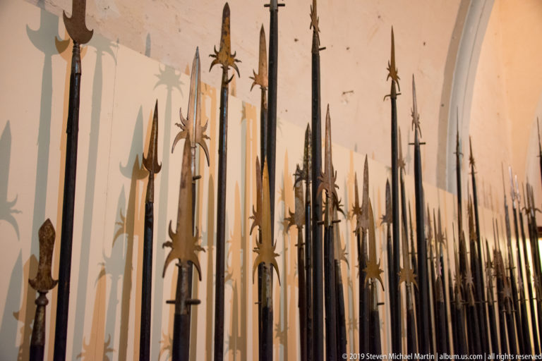 Long rows of vertically displayed polearms with spear, halberd, hook, and cross-shaped heads casting shadows on a pale wall.
