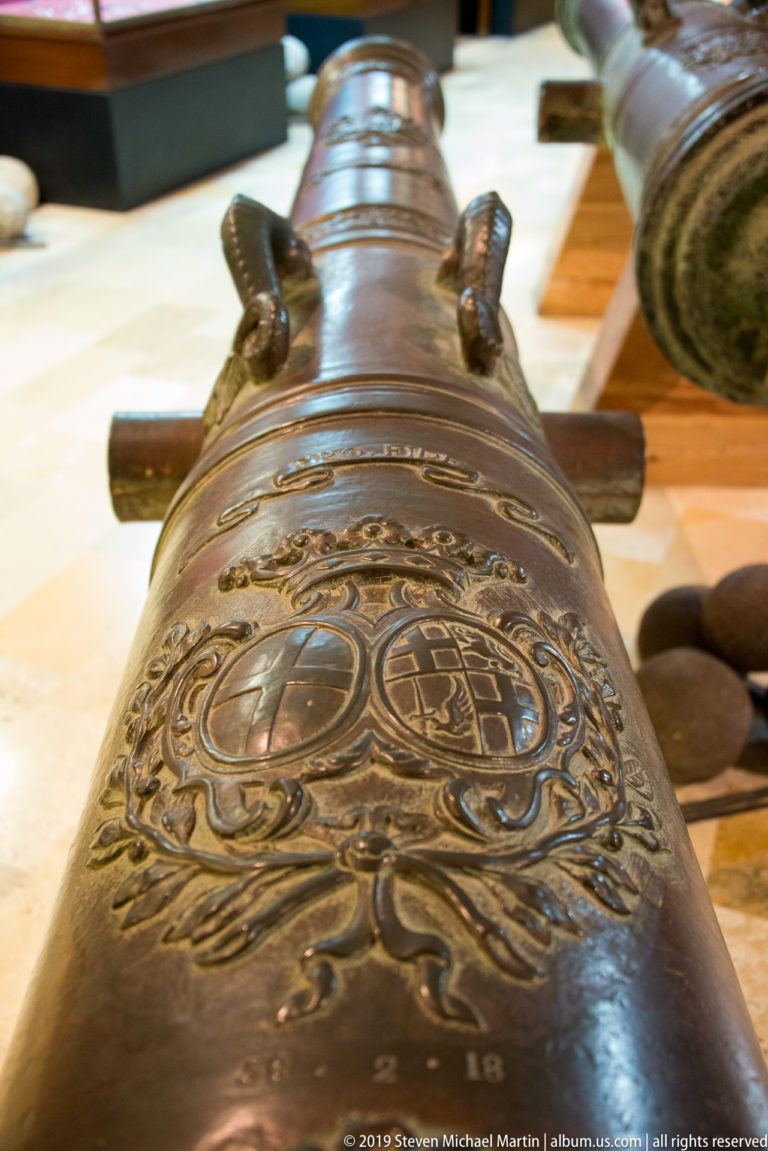 Close view along a bronze cannon barrel decorated with raised heraldic shields, scrollwork, bands, and projecting handles.