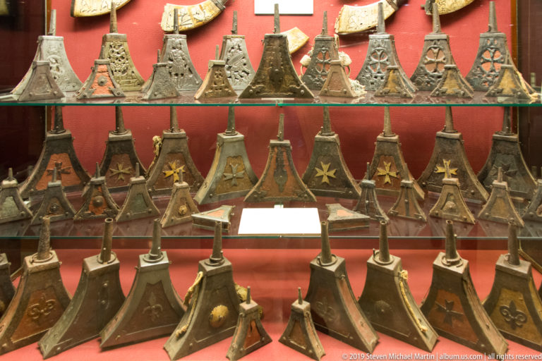 Rows of historic metal polearm heads with varied spear, halberd, and axe-like shapes displayed against red backing.