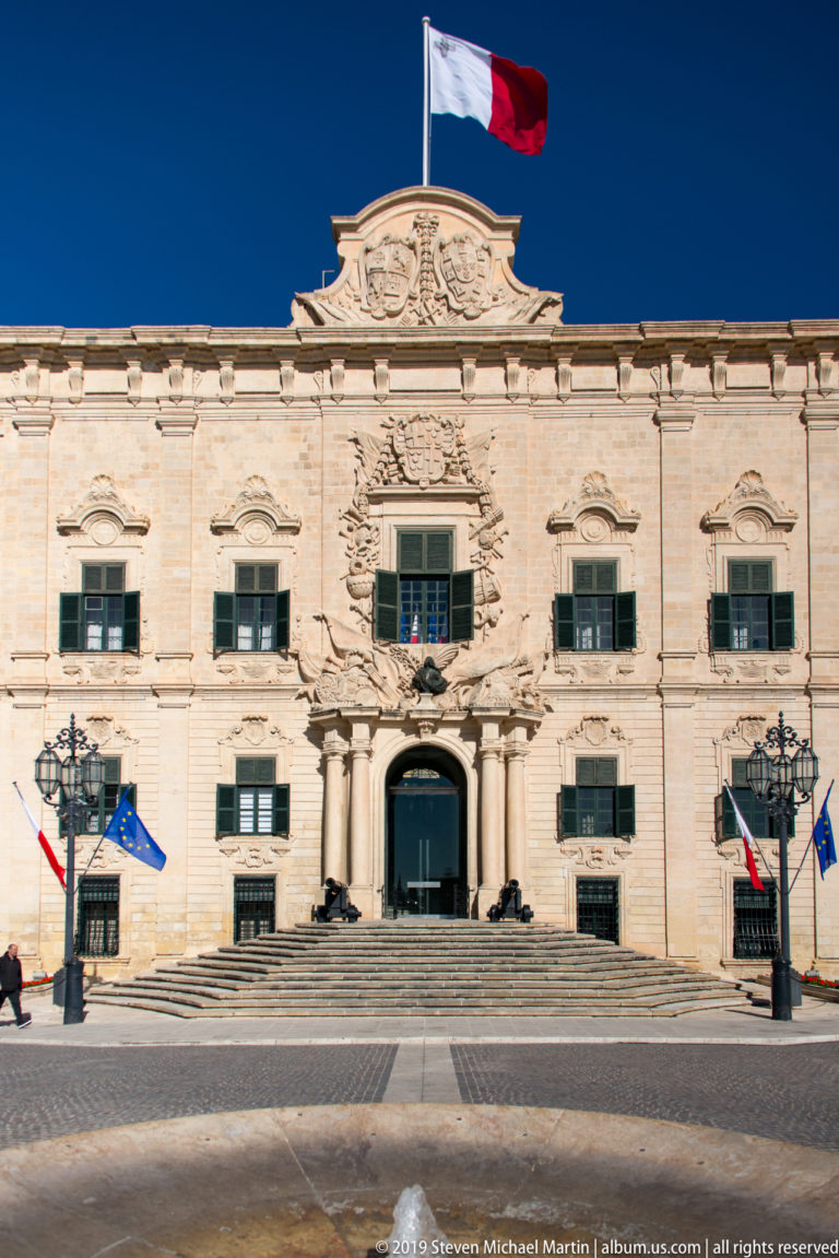 Frontal view of the Auberge de Castille with ornate limestone façade, green shutters, central doorway, Maltese flag, and EU flags.