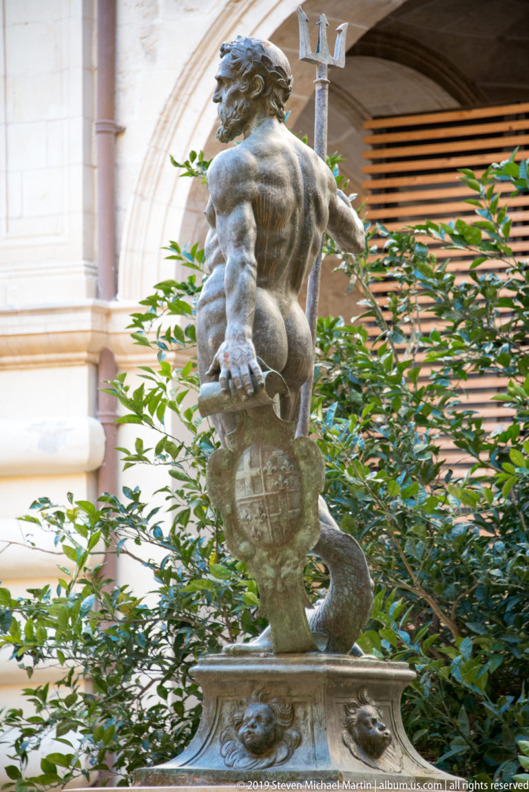 Muscular male statue balancing above a globe-like form and coiling sea creature amid courtyard plants and pale arches.