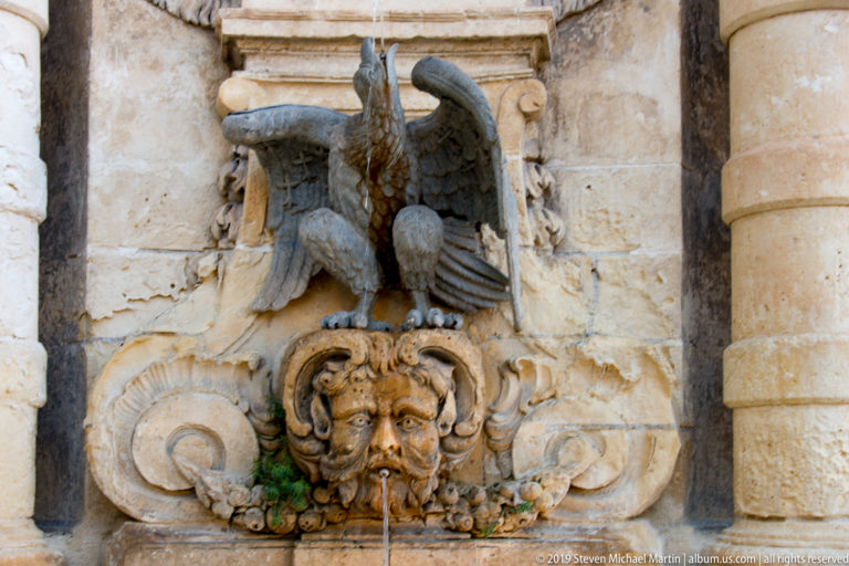 Weathered stone fountain relief with spread-winged eagle above a grotesque mask spout, surrounded by shell scrolls and carved foliage.
