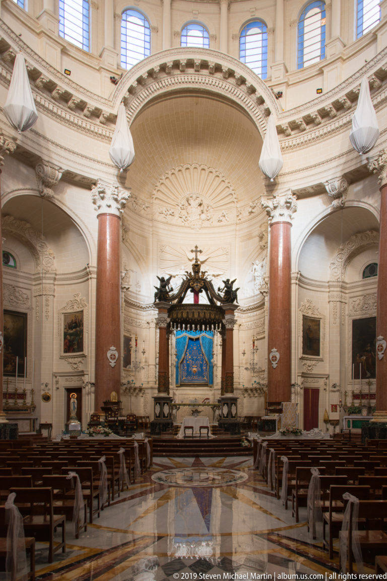 Wide church sanctuary with coffered half-dome, red marble columns, central blue devotional image, side paintings, and polished floor.
