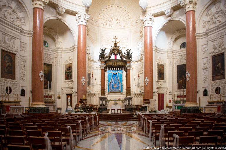 Symmetrical church interior with wooden pews, red marble columns, patterned floor, side paintings, and a blue Marian image above the altar.