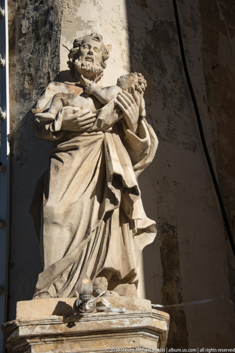 Pale stone statue of a draped male figure holding a cross, lit strongly from one side within a shallow niche.