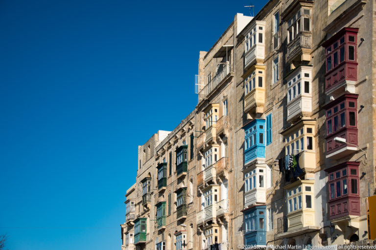 Upward view along a tall limestone façade lined with projecting blue, green, burgundy, and wood-toned enclosed balconies.