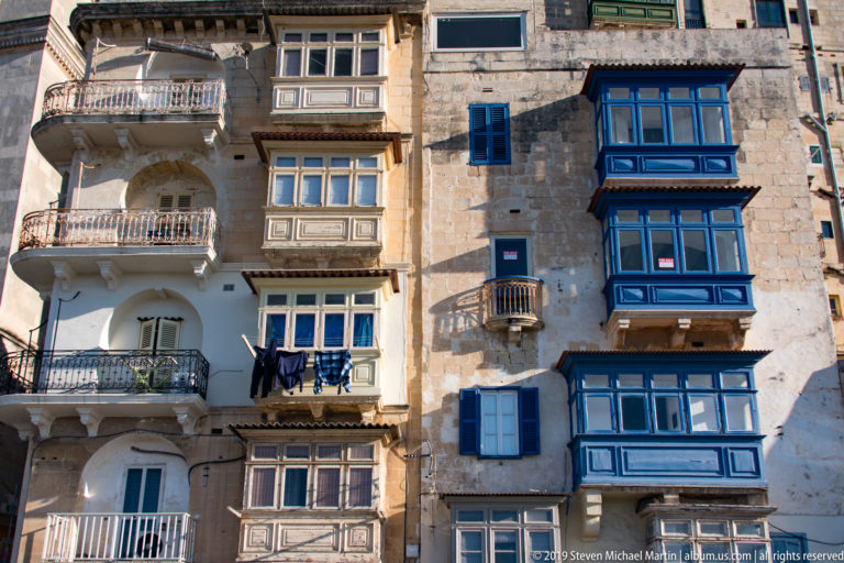 Weathered limestone façades with projecting blue, brown, and white enclosed timber balconies, shutters, railings, and hanging laundry.
