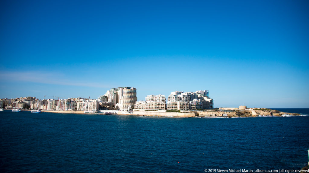 Wide view across deep-blue harbor water toward a dense pale-stone waterfront and rocky shoreline under a clear sky.