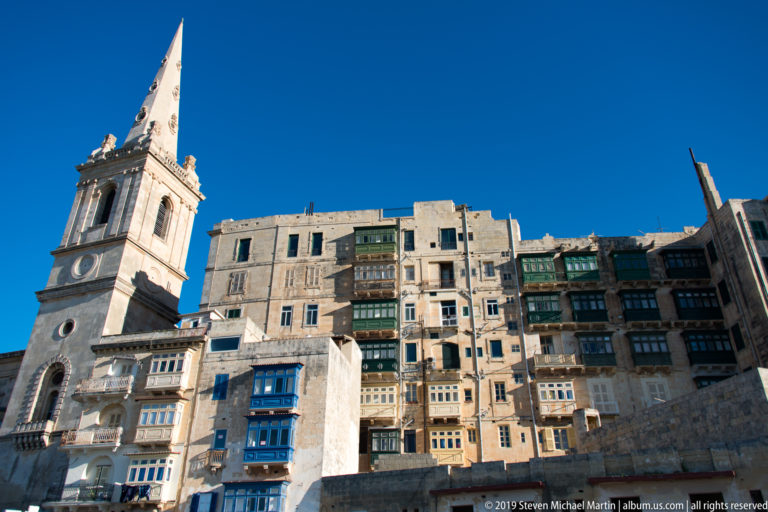 Low-angle view of a pale stone church tower beside tall limestone residences with projecting green and blue balconies.