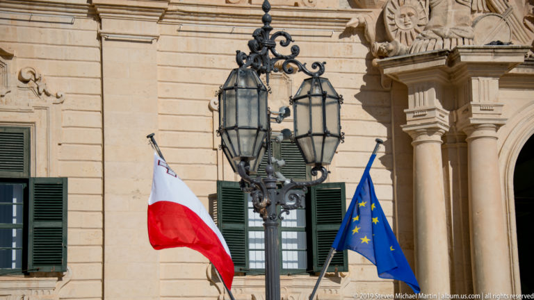 Maltese and European Union flags beside an ornate black lantern on a limestone façade with green shutters and carved decoration.