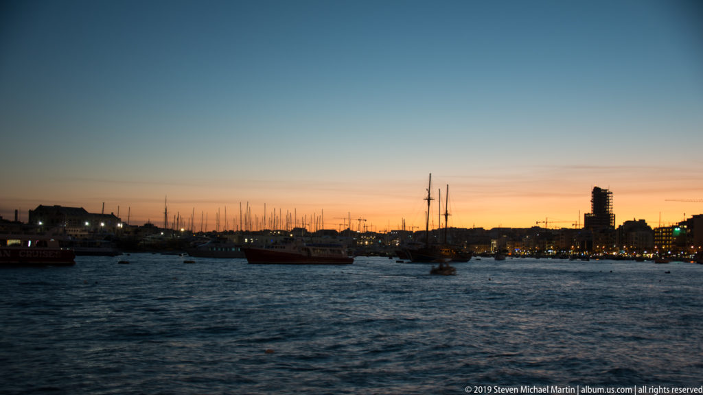 Dusk harbor view with boats and masts on dark water beneath an orange-and-blue sunset sky and low city skyline.