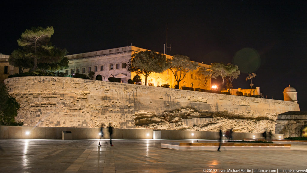 Wide paved plaza at night beside a tall illuminated limestone fortification wall, with small pedestrians, trees, and a building above.