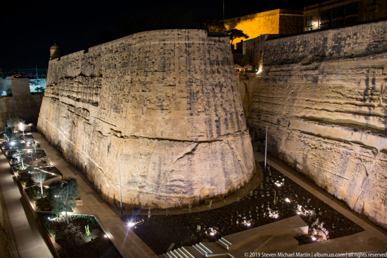 Large rounded limestone bastion illuminated within a deep rock-cut ditch, with sheer walls and landscaped paths at its base.
