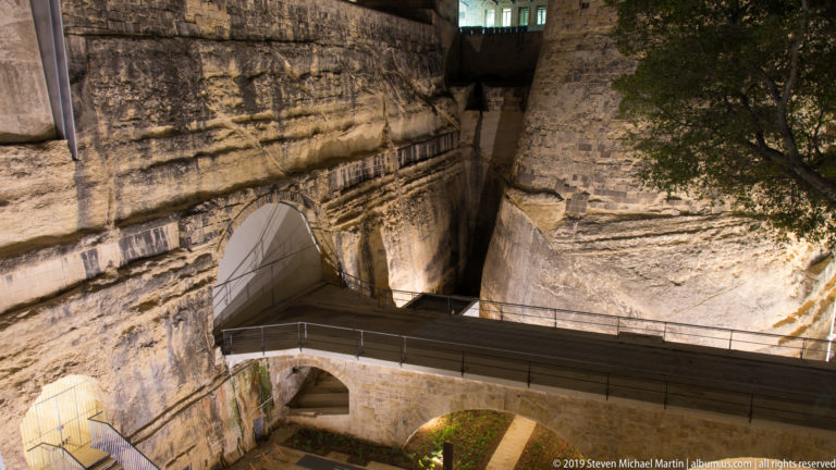 Modern pedestrian bridge crossing between massive limestone walls and arched openings in a deep rock-cut fortification ditch.