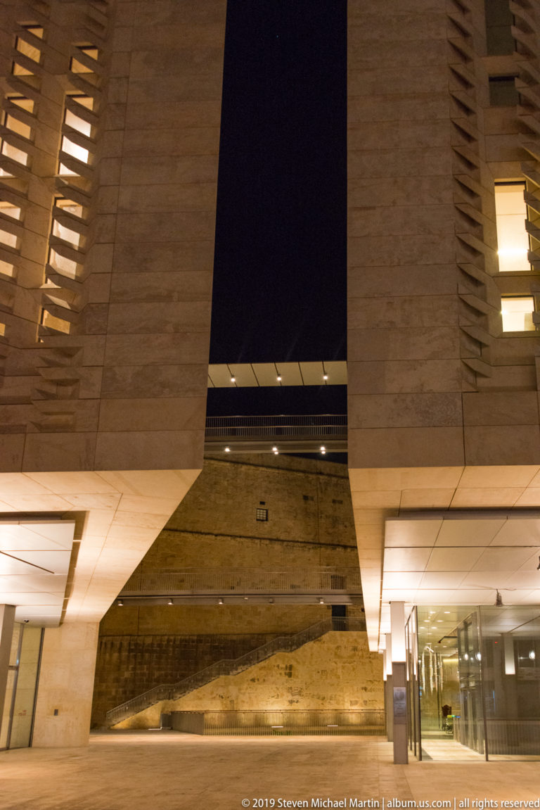 Modern limestone buildings frame an open gap above a rough historic stone wall, with recessed lighting illuminating the preserved masonry below.