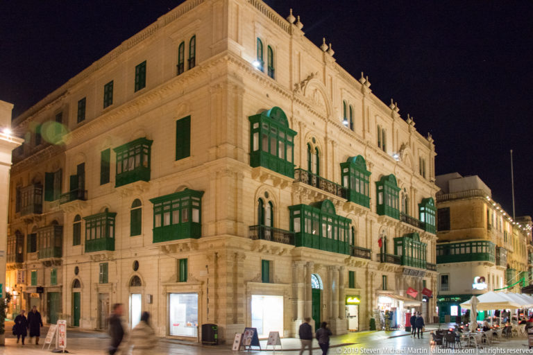 Pale limestone corner building at night with rows of green enclosed timber balconies, ground-floor shops, and pedestrians.