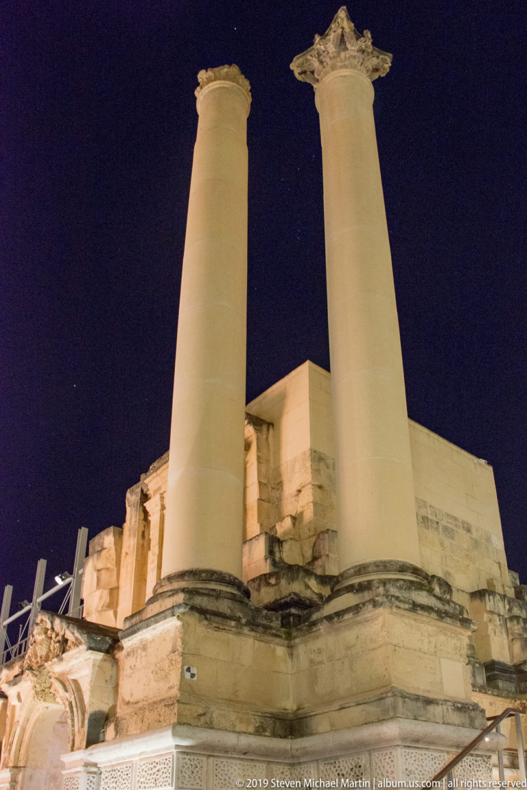 Low-angle night view of two tall pale stone columns with carved capitals rising above fragmented masonry and ruins.