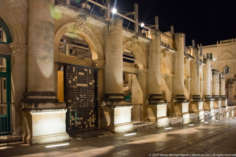 Night view of a weathered stone arcade with thick columns, shallow arches, and a tall iron gate in one bay.
