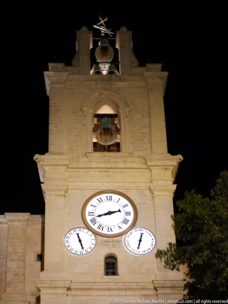 Illuminated square limestone tower with an open bell chamber above three bright clock dials at night.