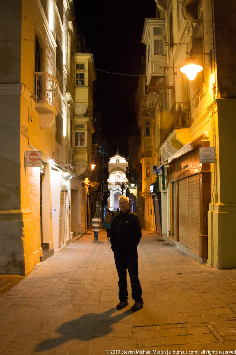 Single pedestrian crossing a warmly lit narrow street at night between shuttered shops and buildings with projecting balconies.