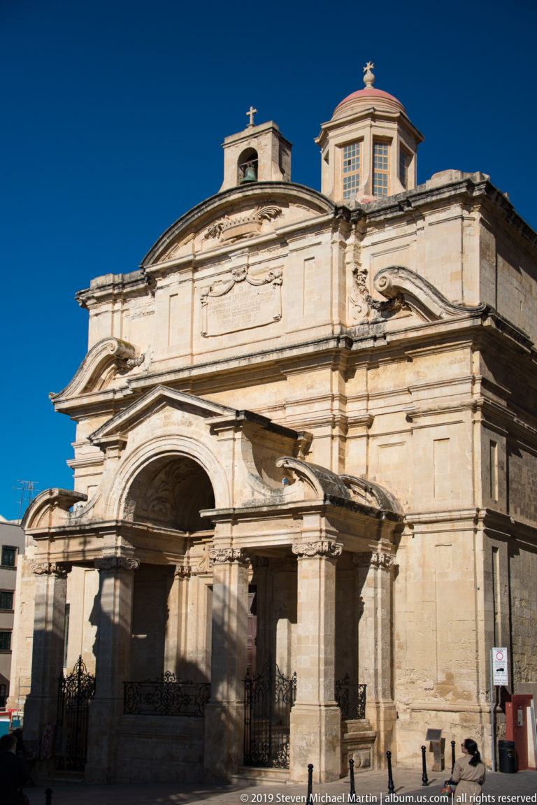 Limestone Baroque church façade with deep arched portico, columns, curved pediments, layered cornices, rooftop lanterns, and crosses.