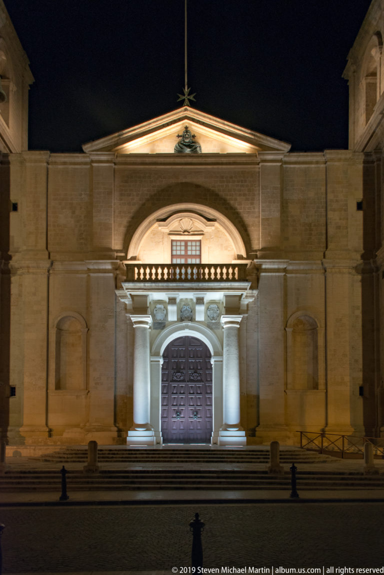 Frontal night view of a limestone church entrance with arched doorway, paired columns, balustrade, and triangular pediment.