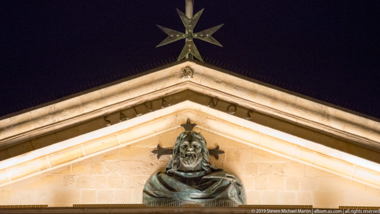 Upward night view of a bronze bust beneath a large eight-pointed Maltese cross mounted at the apex of a stone pediment.