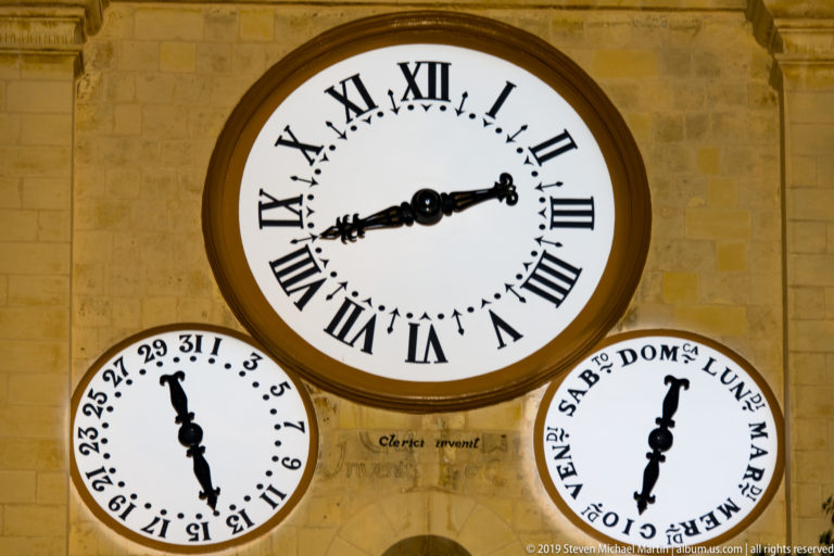 Close view of three illuminated clock dials on limestone, with a large Roman-numeral clock above two smaller circular indicators.