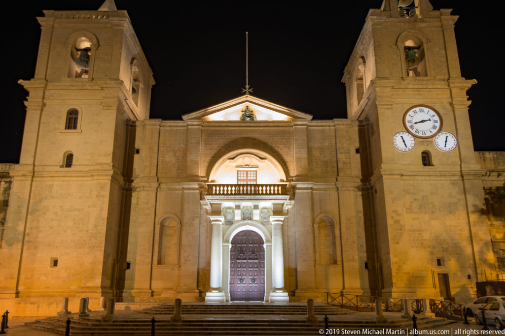 Illuminated limestone church façade at night with a central arched doorway, broad steps, twin bell towers, and multiple clock faces.