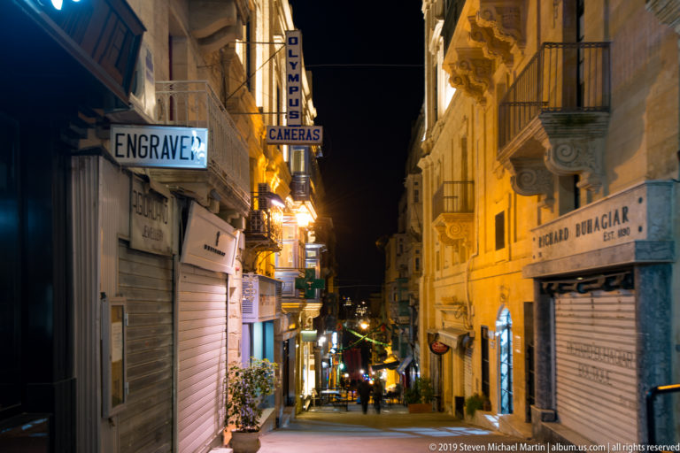 Steep narrow street descending between limestone buildings with projecting balconies, shop signs, shutters, and warm night lighting.