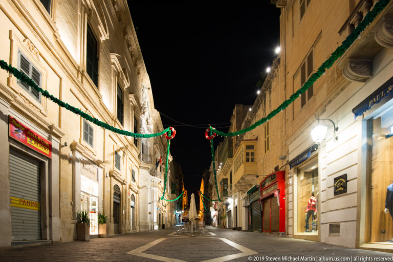 Mostly empty pedestrian shopping street at night with limestone buildings, closed storefronts, and green-and-red overhead decorations.