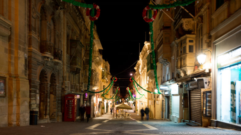Night street with festive garlands, ornate limestone façades, enclosed balconies, shops, pedestrians, and a red telephone box.