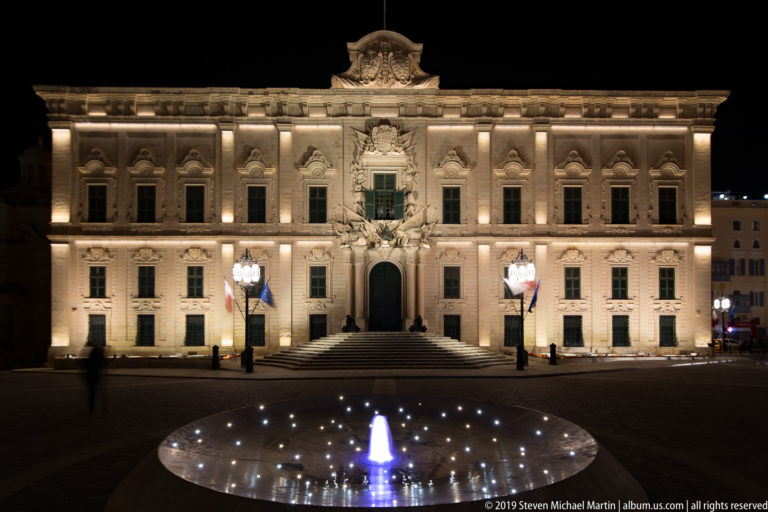 Illuminated Baroque façade of Auberge de Castille with central staircase and ornate crest, seen beyond a circular lit fountain.