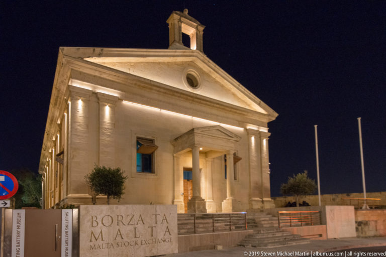 Illuminated Malta Stock Exchange building with triangular pediment, oculus, columned entrance, and inscribed stone forecourt wall.