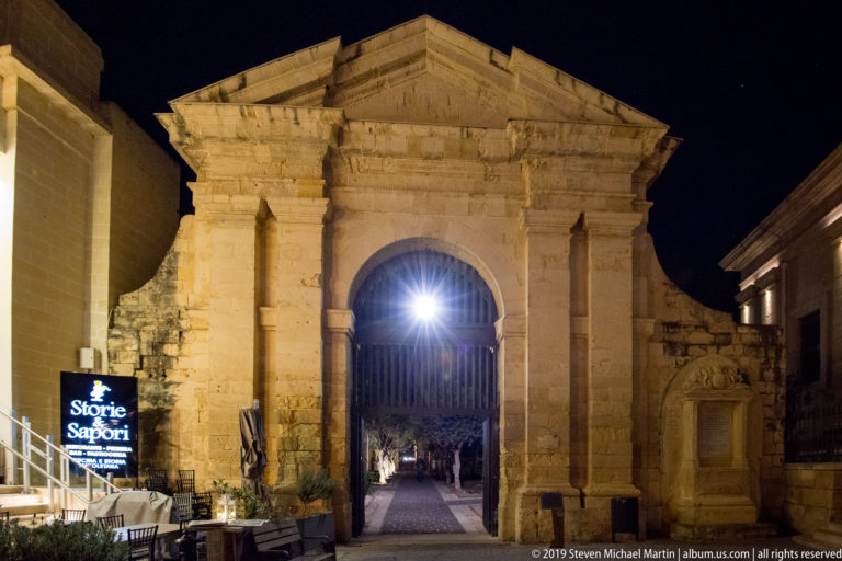 Illuminated monumental limestone gateway with round arch and iron gate opening onto a straight path at night.