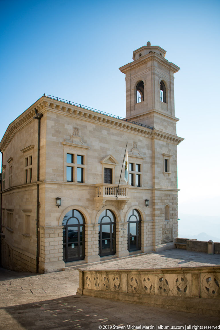 Palazzo Pubblico stone façade with arched entrances, central balcony, San Marino flag, and square civic tower above a paved terrace.