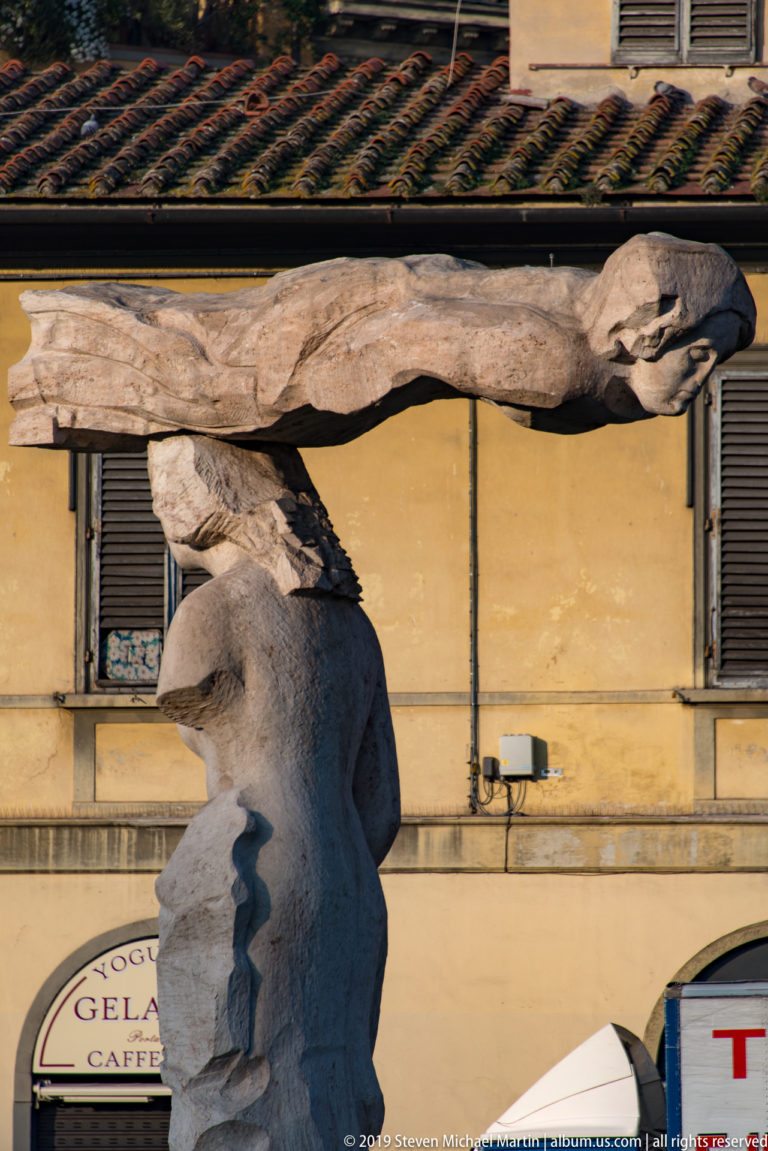 Elongated nude male sculpture extending horizontally above a female figure, with a yellow Florentine façade and tiled roof behind.