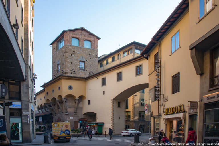 Florence street near Ponte Vecchio with a masonry tower, broad overhead passage, shops, pedestrians, and delivery vehicles.