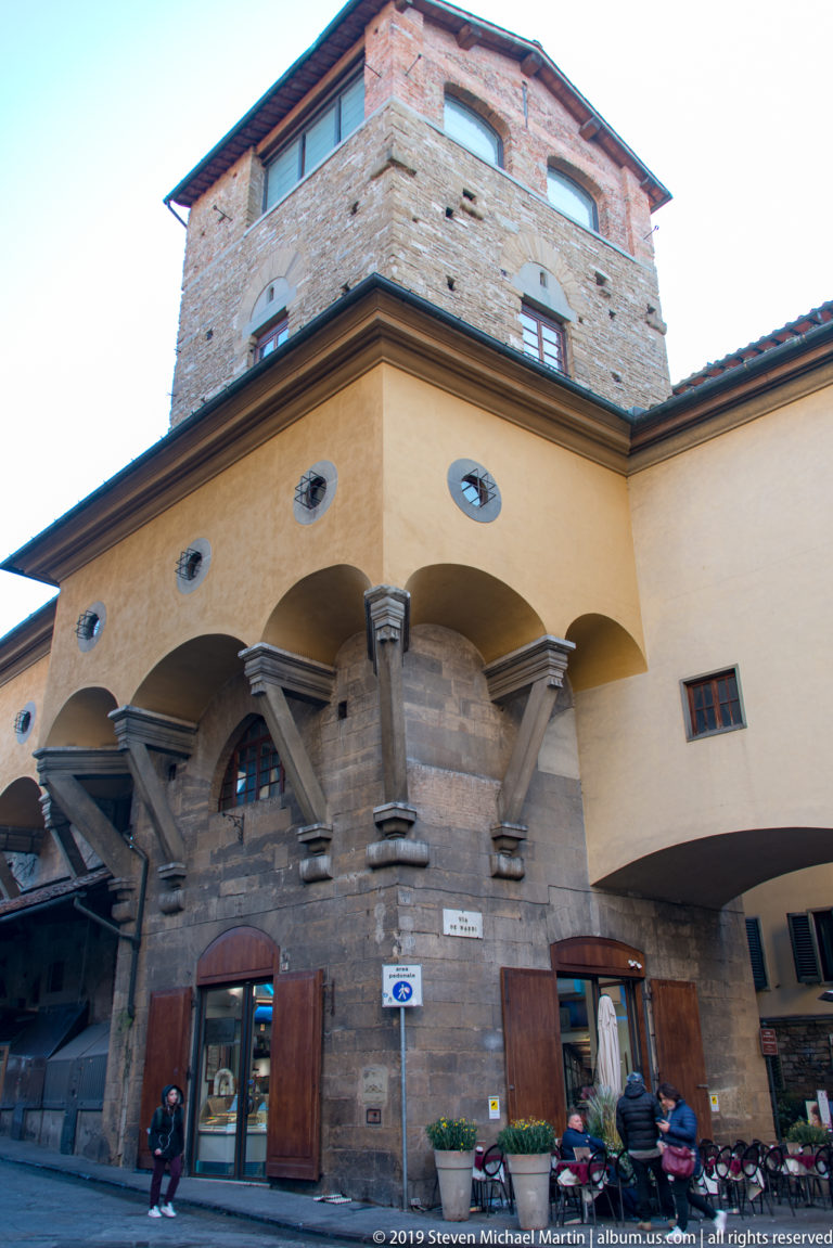 Stone tower rising above a warm plastered arcade with heavy brackets, circular openings, shops, and pedestrians near Ponte Vecchio.