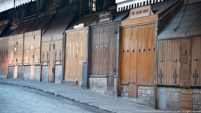 Row of closed wooden Ponte Vecchio shopfronts with iron hinges, studs, locks, and jewelry signs above several doors.