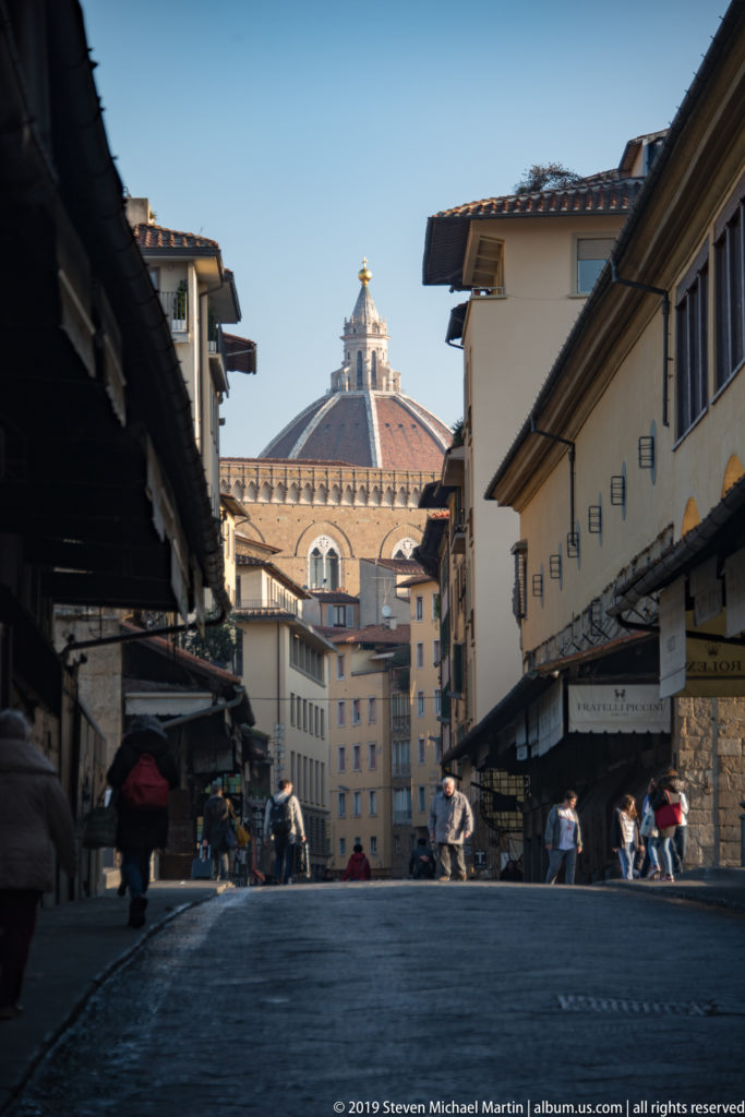 Pedestrian street across Ponte Vecchio lined with closed shopfronts, with Florence Cathedral’s dome rising in the distance.