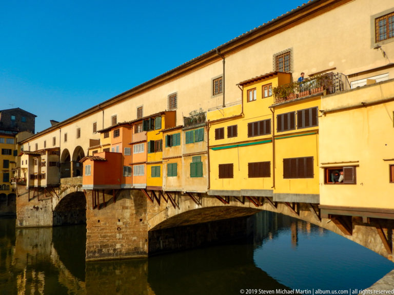 Sunlit yellow, ochre, and salmon shop rooms projecting from Ponte Vecchio above a large stone arch.