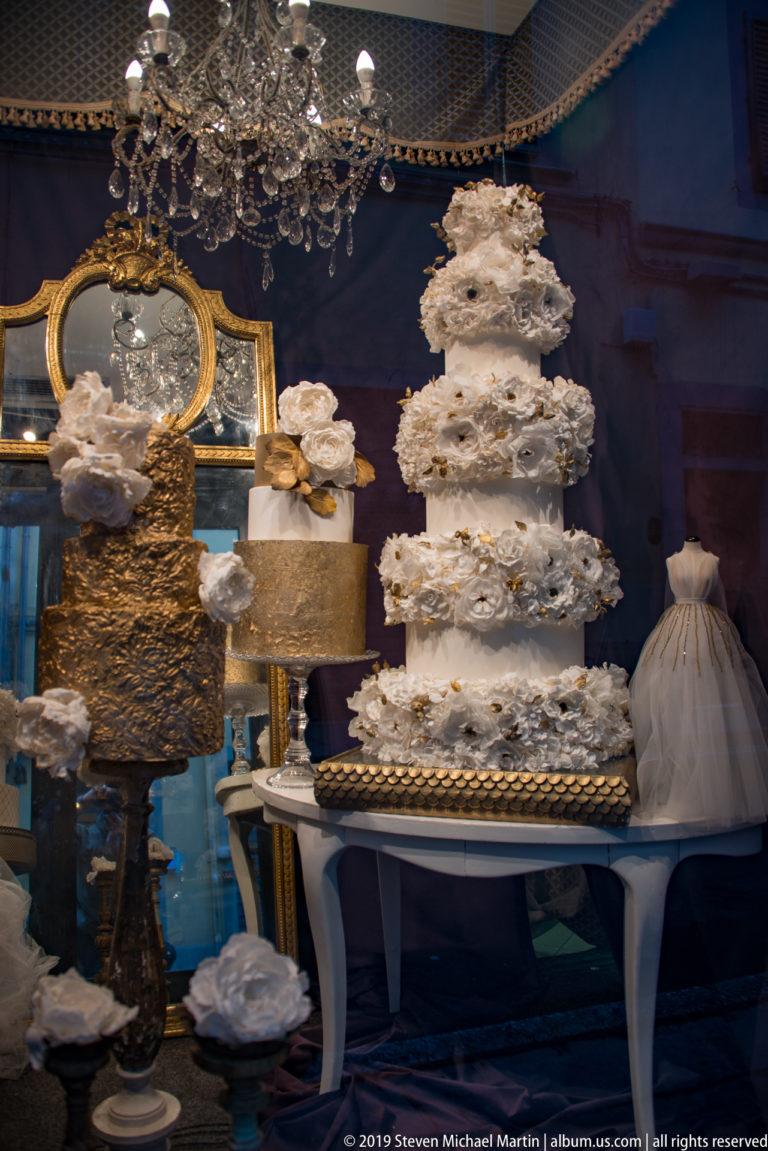 Ornate shop interior with tall white tiered decorations, gilded details, sculptural objects, and a large mirror against a dark blue wall.